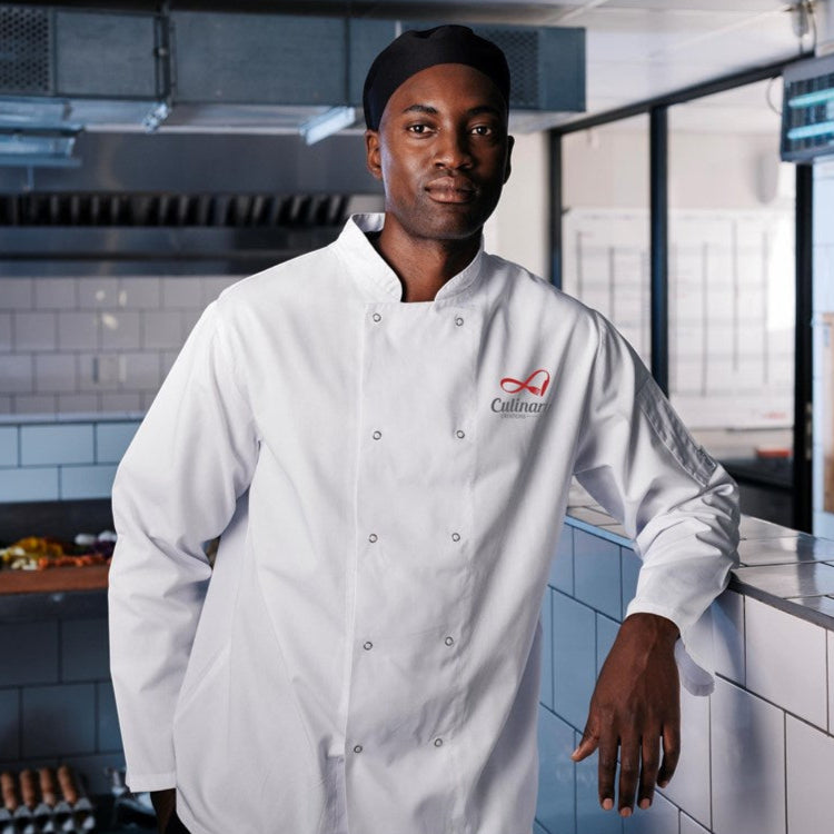 A chef wearing a long-sleeved standing in a kitchen leaning on a counter facing forward