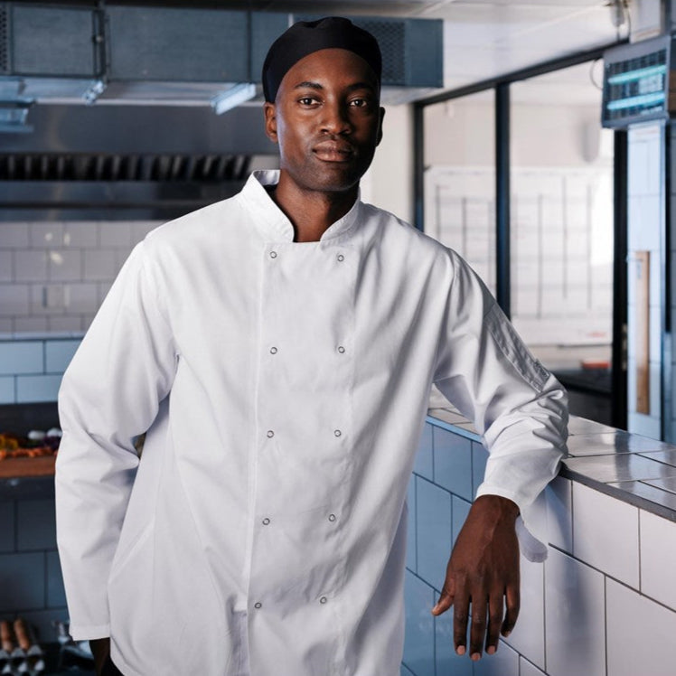 A chef wearing a long-sleeved standing in a kitchen leaning on a counter facing forward
